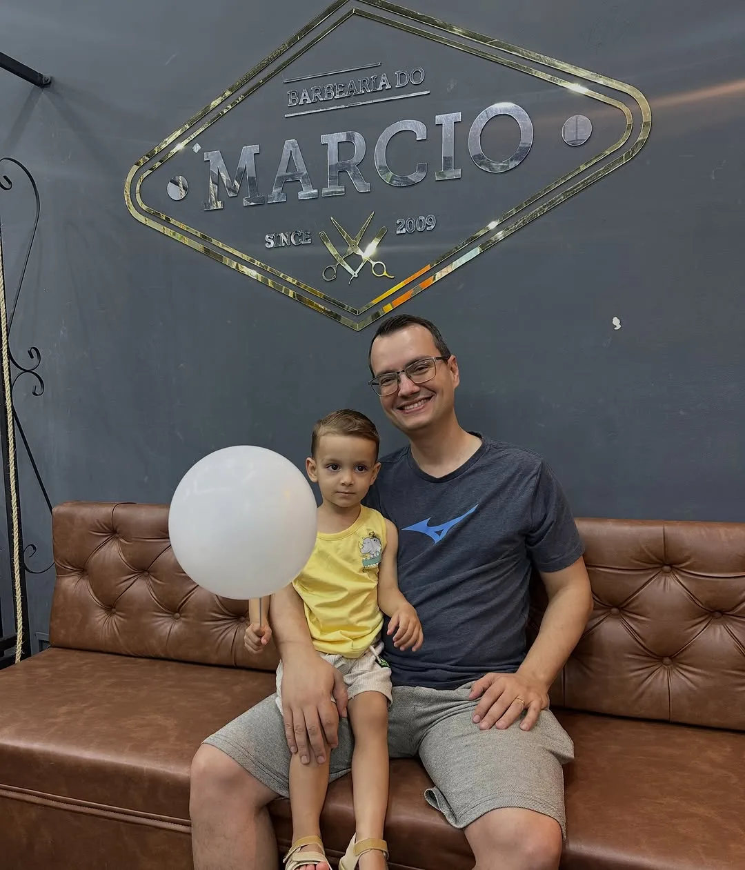 Cliente e criança no sofá de couro, sob o letreiro da barbearia