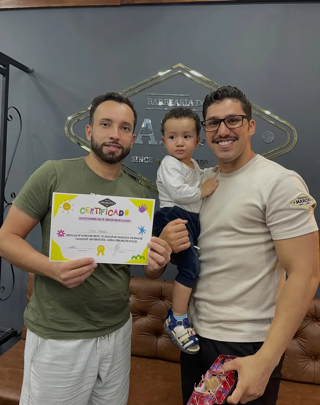 Pai e barbeiro com o bebê e o certificado do primeiro corte